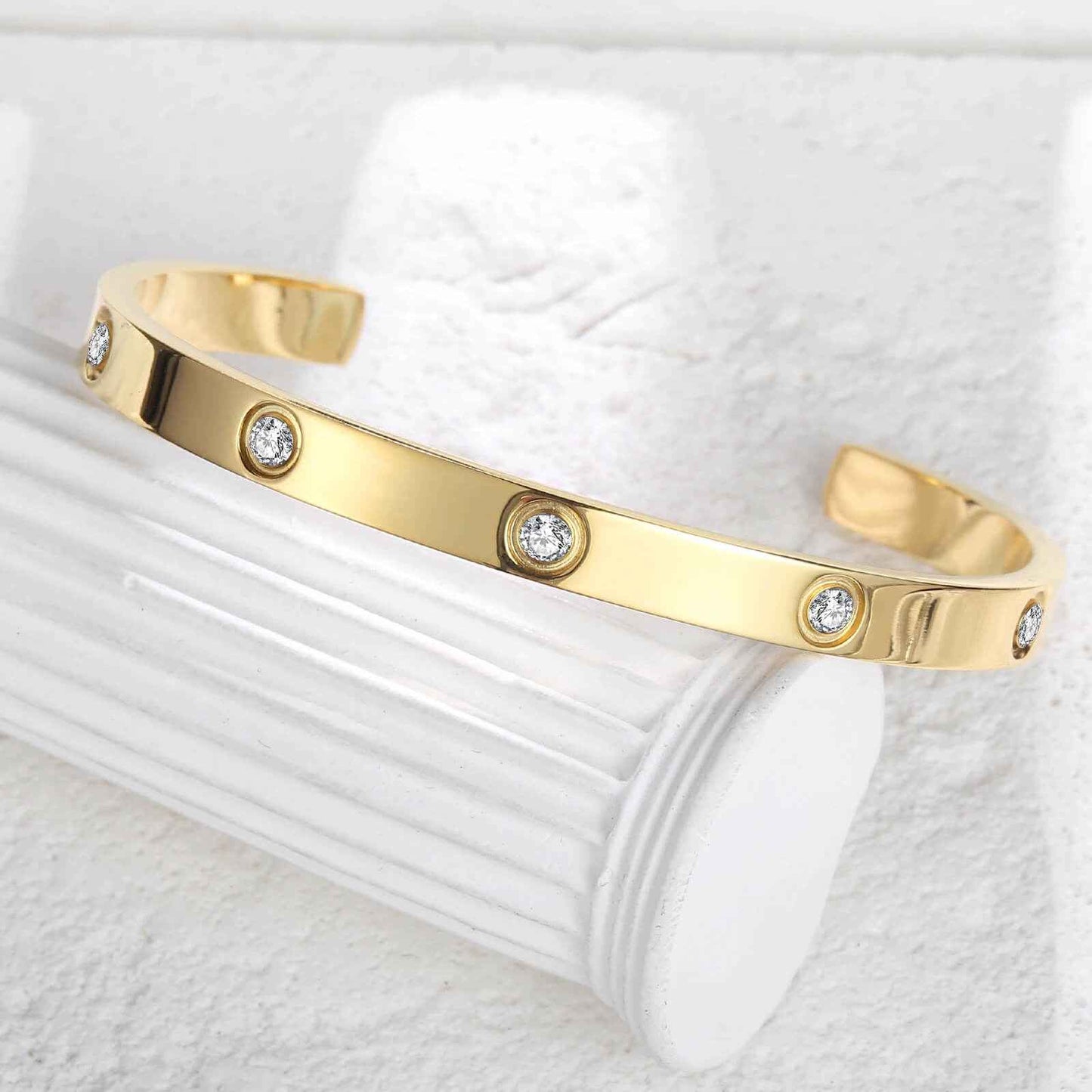 Gold love cuff bracelet displayed on white column, featuring high-polished finish and embedded zirconia for a luxurious look.