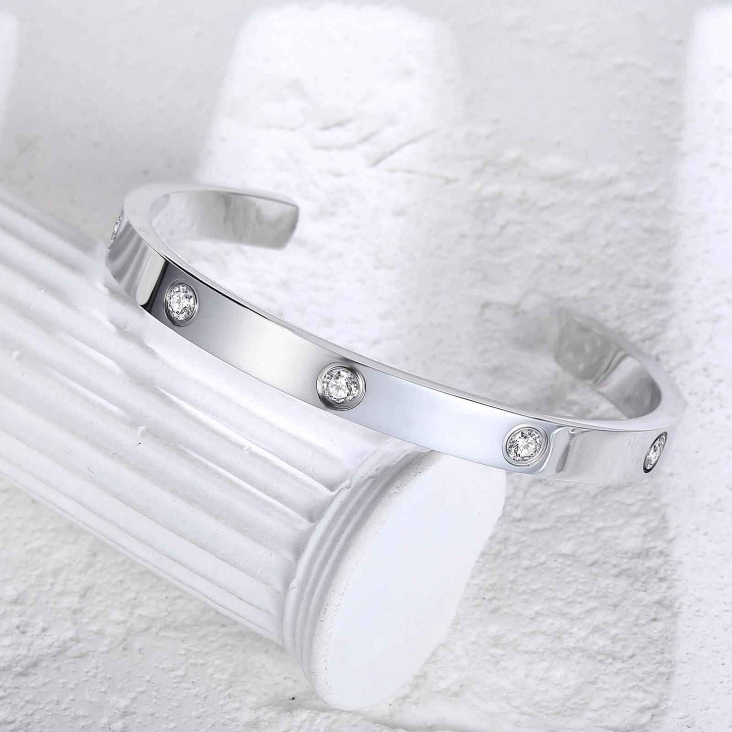 Silver love cuff bracelet resting on white column, showcasing its minimalist waterproof design with sparkling zirconia accents.