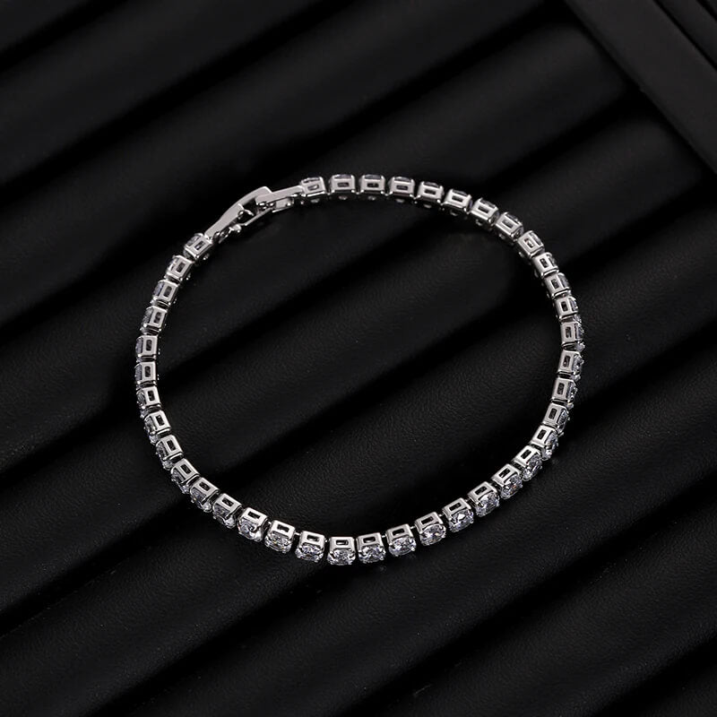 Silver iced out tennis bracelet with clear zirconia laid flat on ribbed black surface, stylish hip hop jewelry piece.
