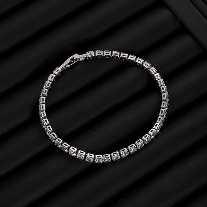 Silver iced out tennis bracelet with clear zirconia laid flat on ribbed black surface, stylish hip hop jewelry piece.